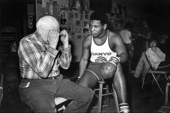 Mike Tyson ao lado de Cus D’Amato, seu treinador, recebendo instruções técnicas e mentais durante o treinamento. A imagem representa a importância da mentoria, do desenvolvimento psicológico e da construção de mentalidade vencedora como base para a excelência esportiva.