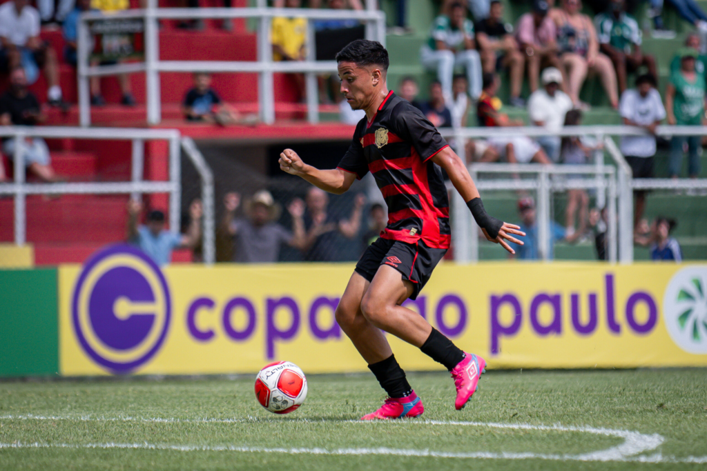Atleta em ação durante partida da Copa São Paulo, executando um drible com a torcida ao fundo e o logo oficial da competição em destaque. A imagem reforça tomada de decisão, coragem ofensiva e pressão competitiva em torneios de alto nível do futebol de base.