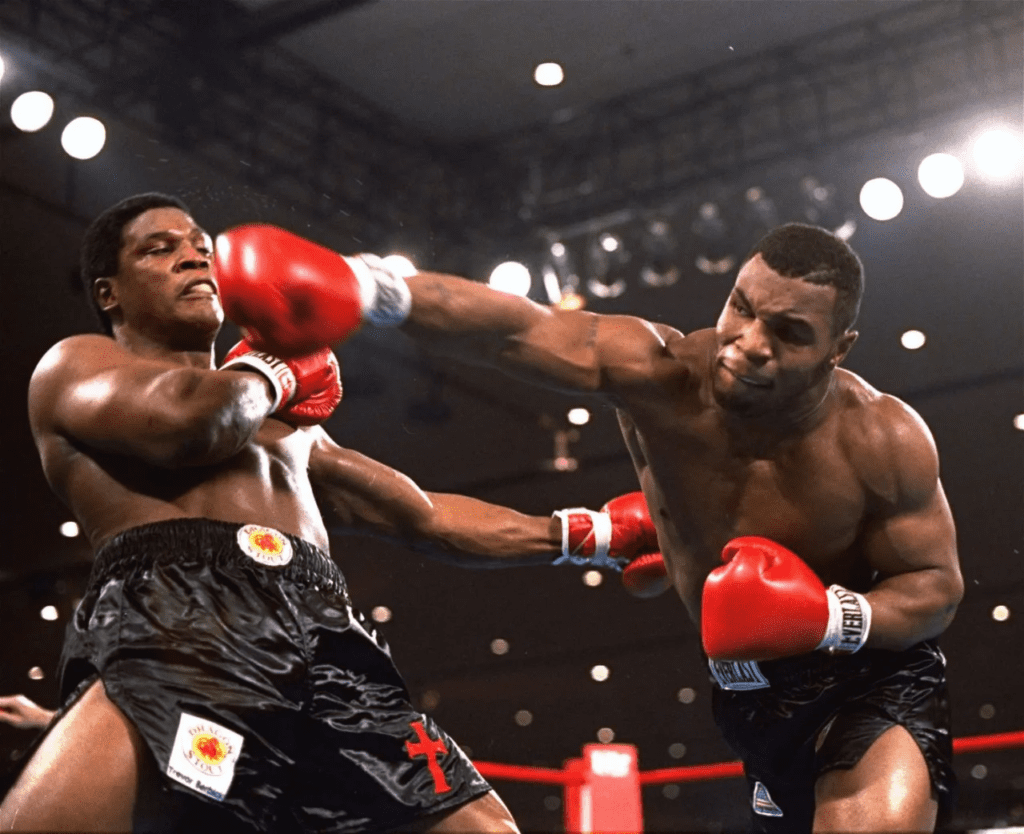 Mike Tyson executando um jab direto com força total, enquanto o adversário tenta se esquivar dentro do ringue. A imagem reforça precisão técnica, leitura corporal do oponente e sincronização entre mente e corpo, fundamentais para a performance no boxe profissional.