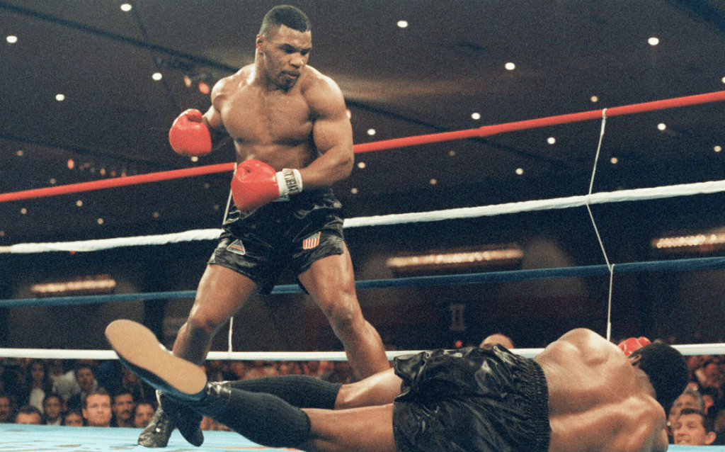 Momento icônico de Mike Tyson aplicando um nocaute, com o adversário caído no chão e a plateia atenta ao impacto da ação. A imagem simboliza agressividade controlada, tomada de decisão rápida, confiança extrema e execução perfeita sob pressão no esporte de alto rendimento.